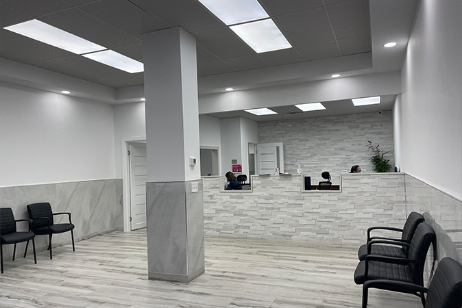 The image shows an interior view of a waiting area within a medical facility, featuring a reception desk with chairs and a large window with blinds.