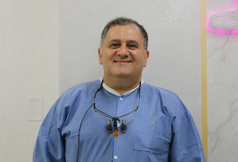 The image is a photograph of a man standing in front of a wall with a pink object on it, wearing glasses and a blue shirt. He has dark hair and appears to be smiling slightly.