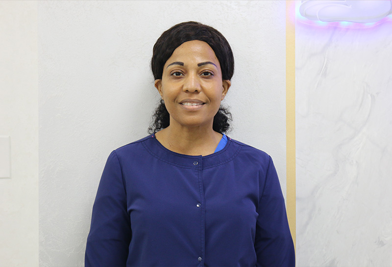 The image shows a woman standing against a wall, wearing a blue top and smiling at the camera.
