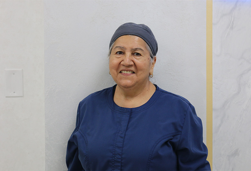 The image features a person standing in front of a wall, smiling at the camera. They are wearing a blue uniform, which suggests they may be a healthcare professional such as a nurse or medical assistant. There is no visible text within the image to provide additional context.
