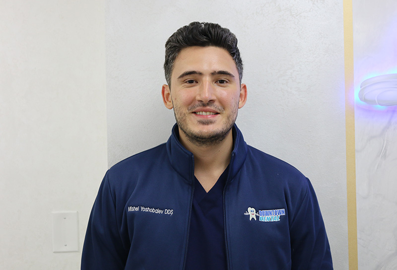 The image is a portrait of a man wearing a blue jacket with a name tag, standing in front of a wall with a graphic design.