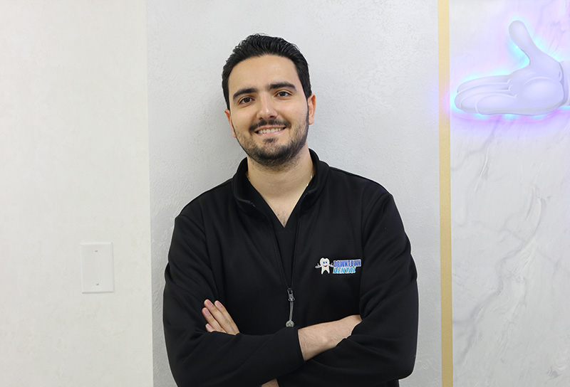The image shows a man standing indoors, wearing a dark jacket and smiling at the camera. He has his arms crossed and is positioned in front of a marble wall with a purple logo on it.