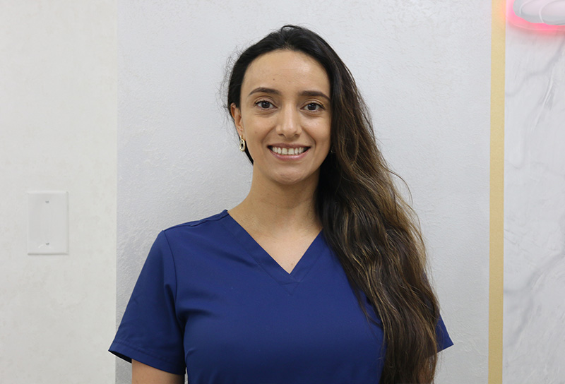 A woman in a blue scrub top stands against a wall, smiling slightly.
