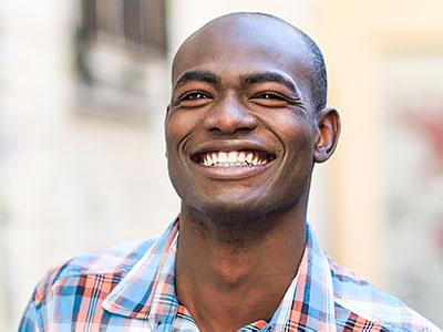 The image shows a smiling man with short hair, wearing a blue and white plaid shirt. He appears to be outdoors during the daytime.