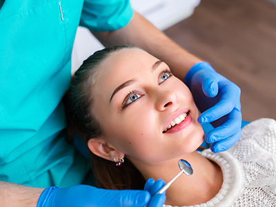 A dental hygienist is performing a cleaning procedure on a patient's teeth, with the patient smiling and wearing glasses.